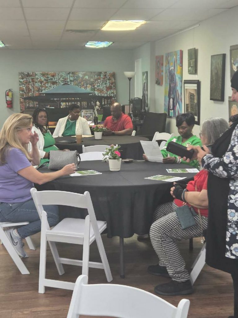 People sitting around tables in an art-filled room, engaged in conversation and looking at papers, with potted plants on the tables.