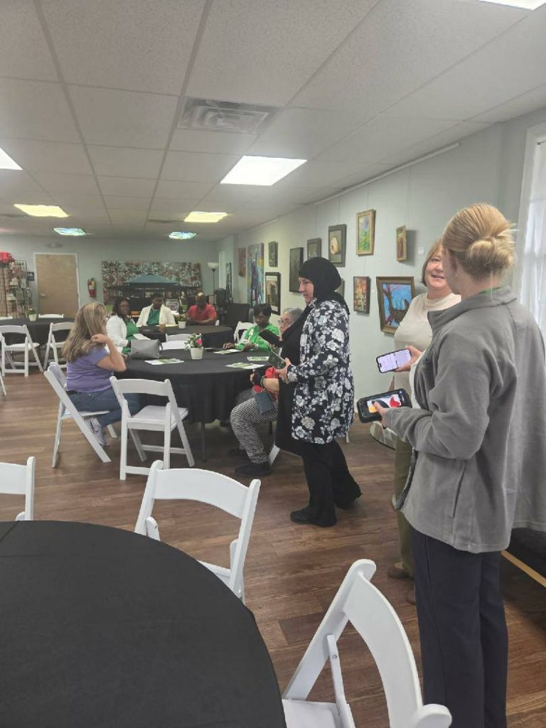 People gathered in a room with round tables, some seated and some standing, conversing. Artwork decorates the walls and natural light enters through windows.