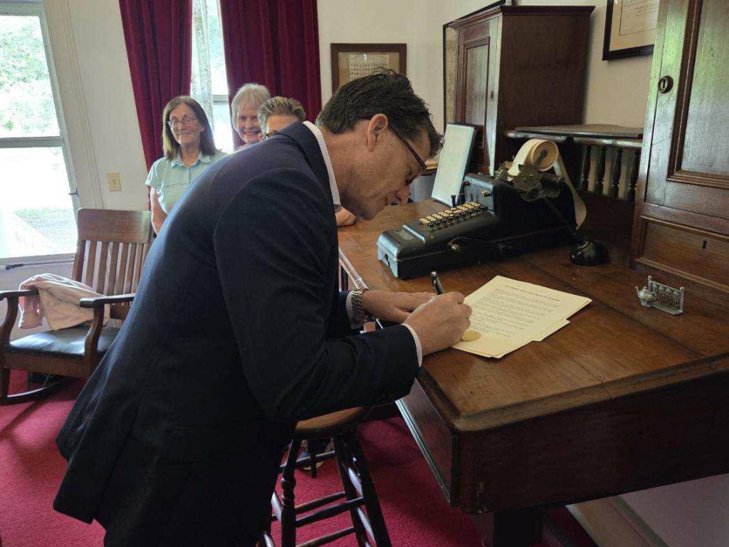 A man in a suit signs documents at a wooden desk while three women stand smiling in the background.