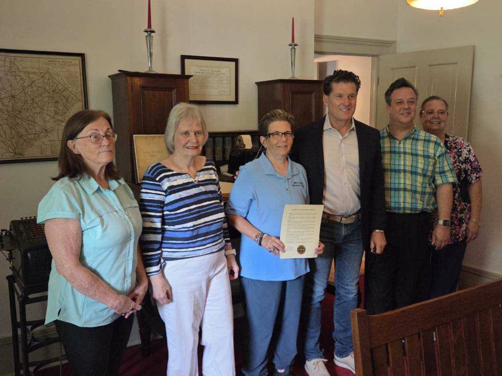Six people pose indoors, one holding an official document, in a room decorated with vintage maps, certificates, and tall red candles.