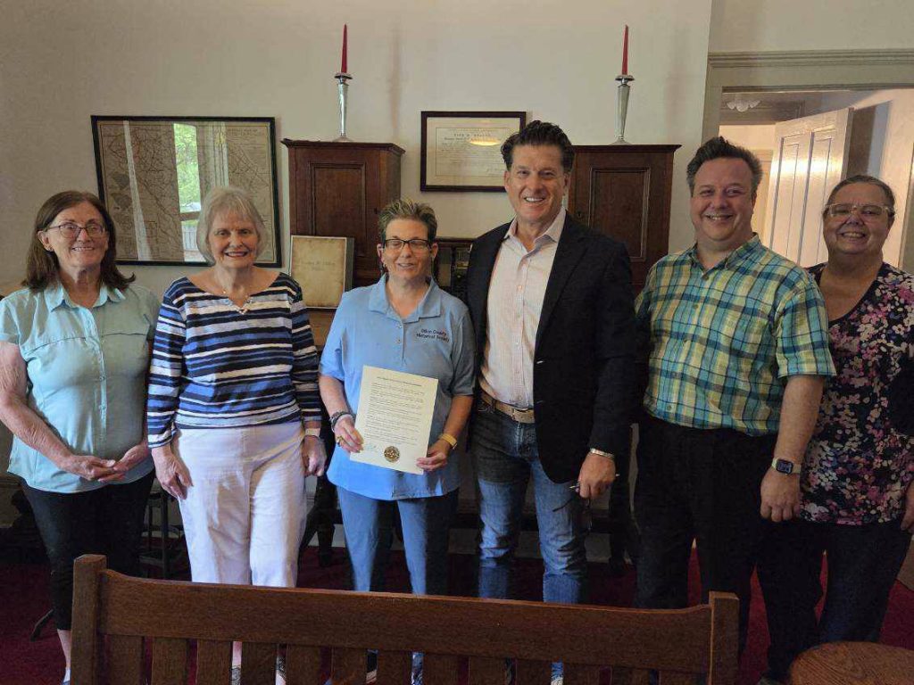 Six adults stand indoors, one holding a certificate. They are smiling in a warmly lit room with wooden furniture, framed maps, and red candles in the background.