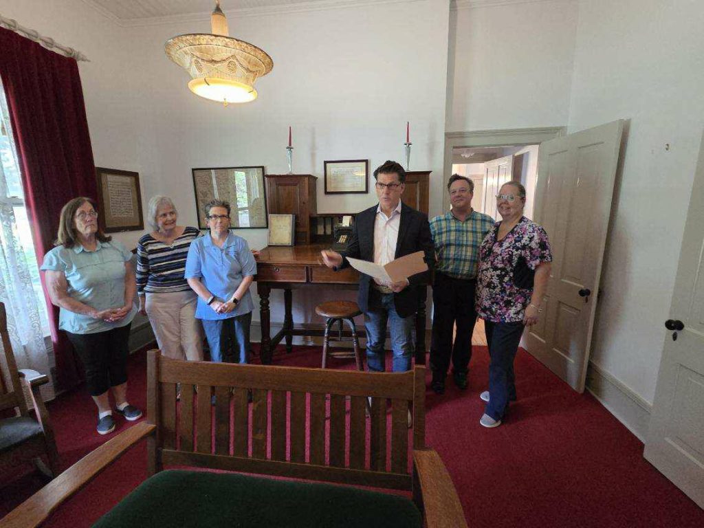 Six people standing in a room with red carpet, framed pictures, wooden furniture, and a man holding papers, suggesting a group meeting or tour.