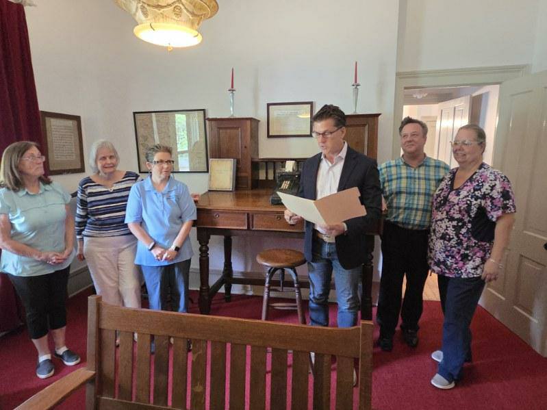 Six adults stand in a room with red carpet and vintage furniture, one man reading from a folder while others watch and smile.