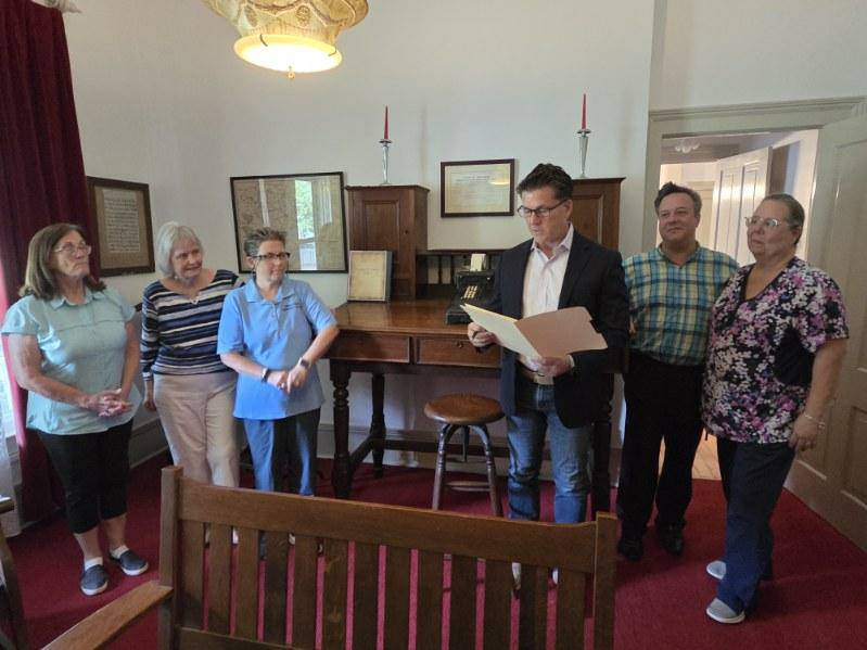 Six adults stand in a small room with vintage decor; one man reads from a folder, while the others listen.
