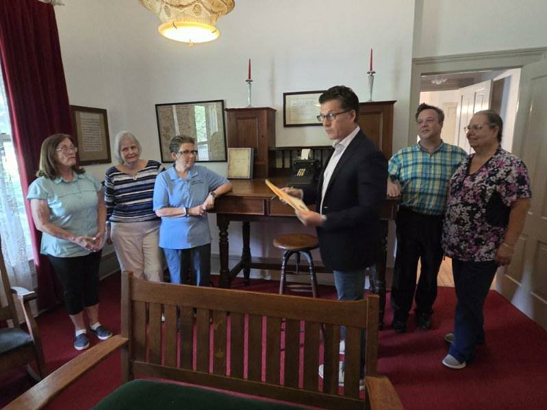 Six adults gather in a historical room with vintage furniture, documents, and decor. One person holds papers and appears to be speaking or presenting to the group.
