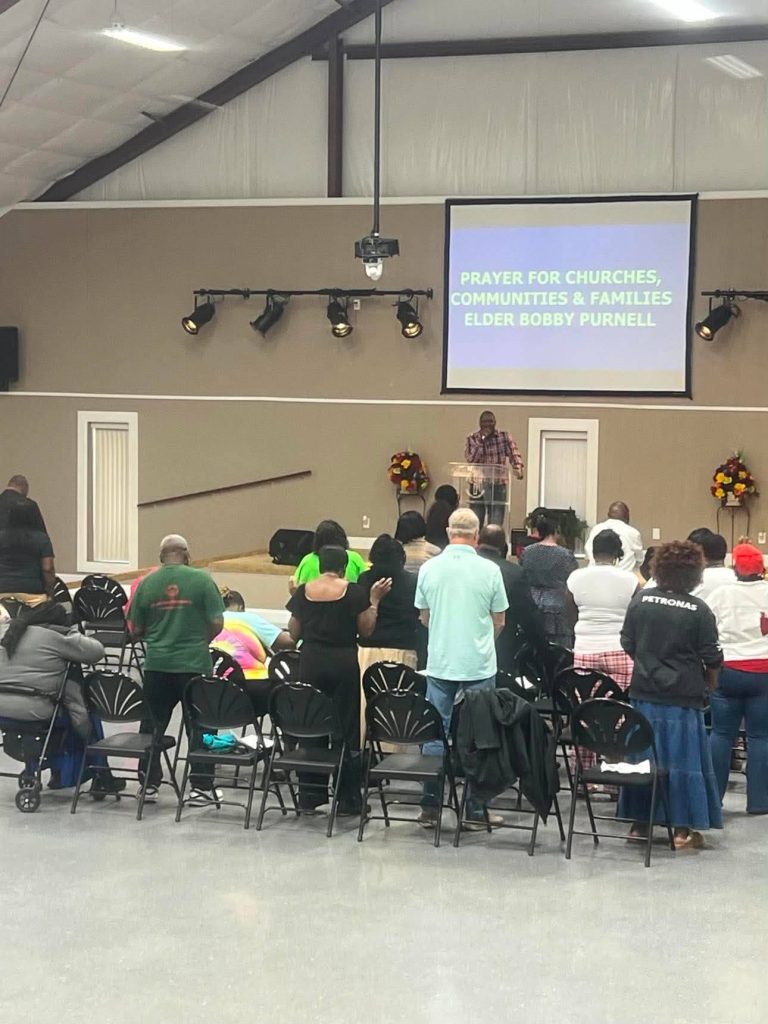 A group of people standing and praying in a church, facing a stage where a speaker presents under a screen with a prayer announcement.
