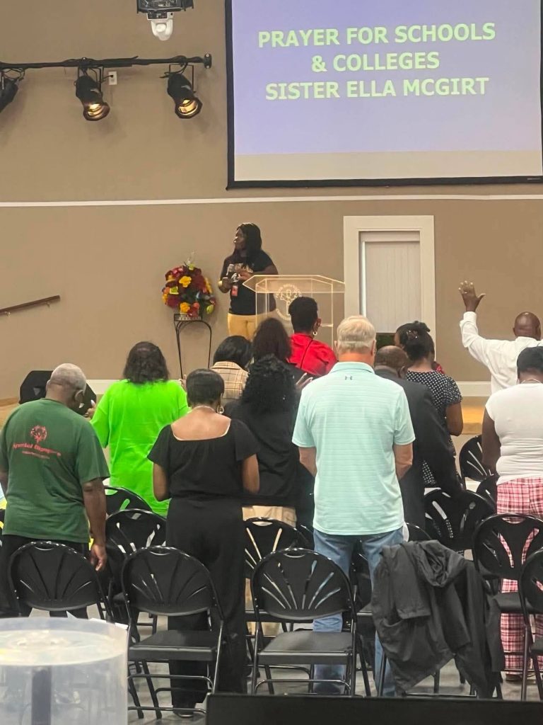A woman speaks at a podium in a church as people stand and pray. Projector screen reads, "Prayer for Schools & Colleges Sister Ella McGirt."