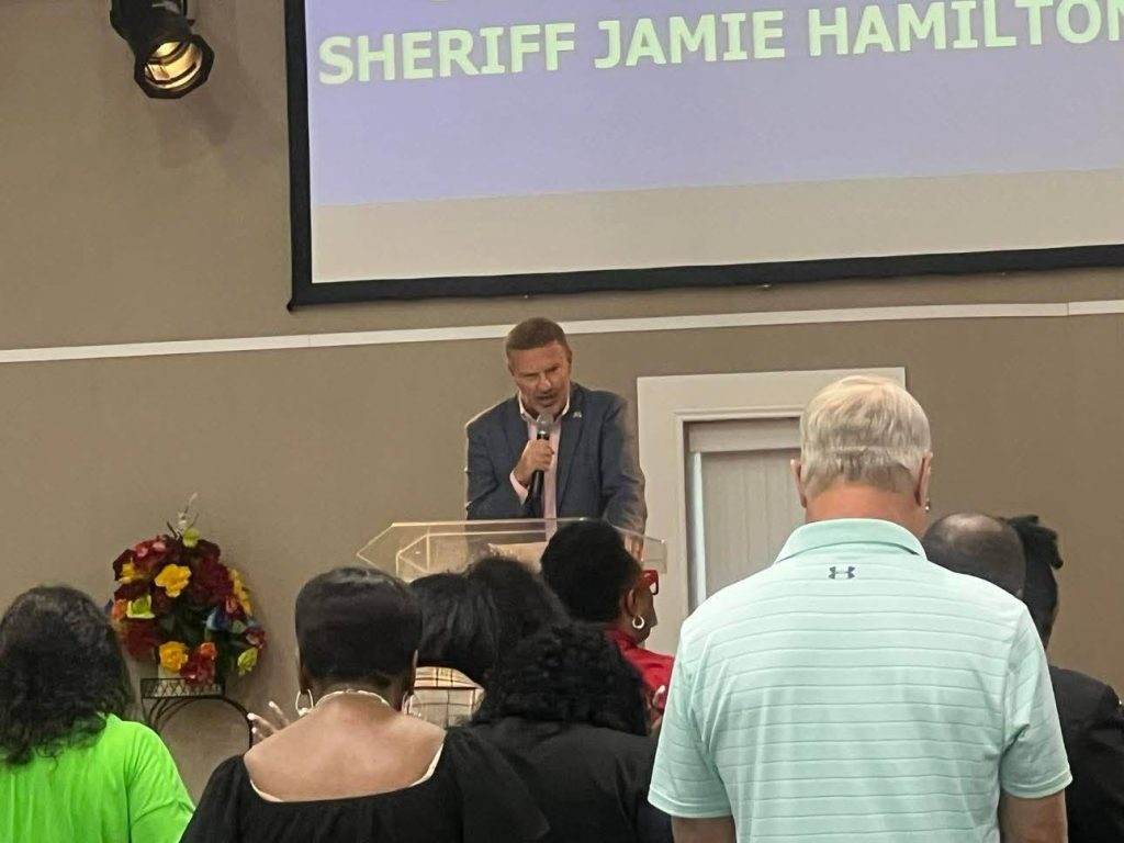 A man speaking at a podium in front of an audience, with a screen behind displaying the words "Sheriff Jamie Hamilton."