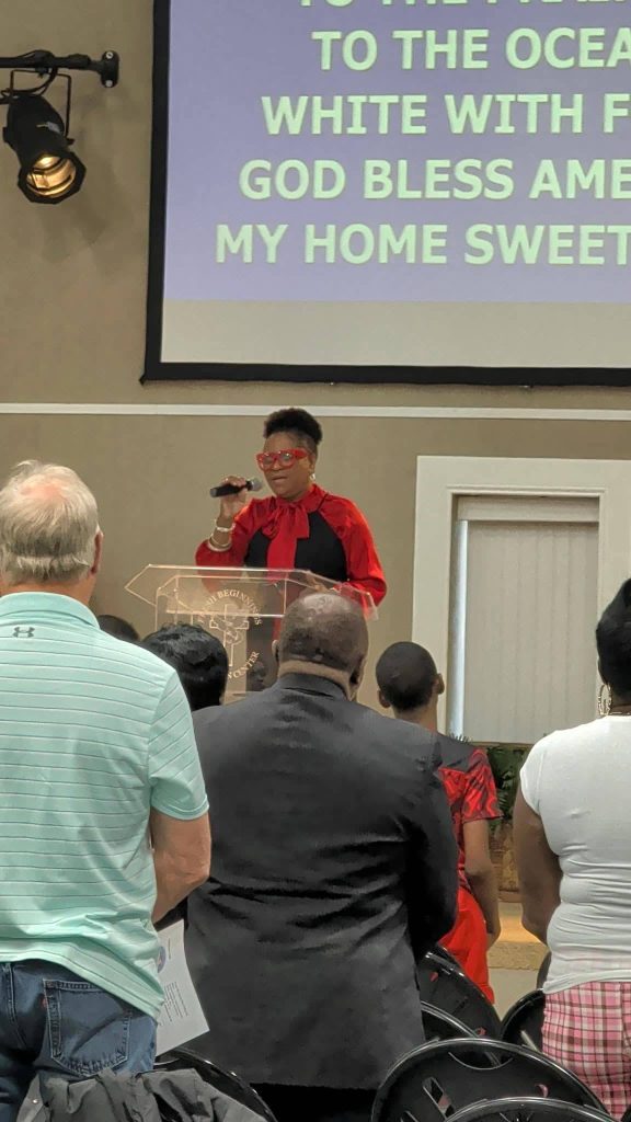 A woman in red and black speaks at a podium to an audience, with song lyrics projected on a screen behind her in a church or event setting.