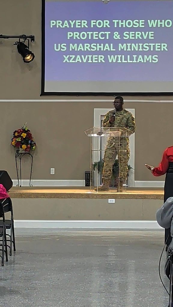 A man in military uniform speaks at a clear podium in a church, with a screen behind reading “PRAYER FOR THOSE WHO PROTECT & SERVE US MARSHAL MINISTER XZAVIER WILLIAMS.”
