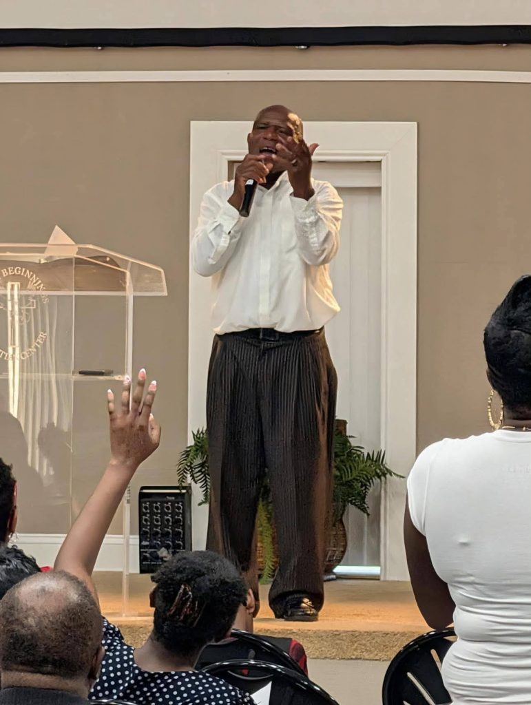 A man in a white shirt sings or speaks into a microphone on stage, while audience members listen and one raises their hand.