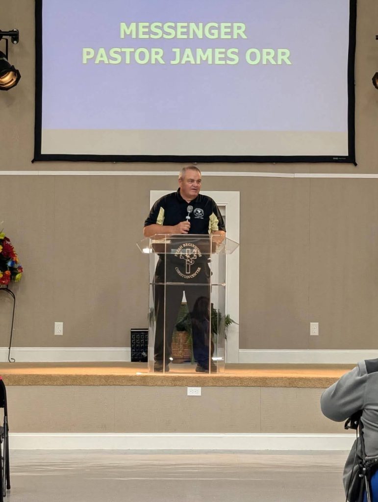 A man stands at a clear podium with a microphone, giving a speech in a church. The screen behind reads "MESSENGER PASTOR JAMES ORR."