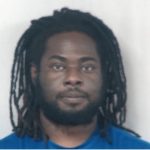 A man with long dreadlocks and a beard, wearing a blue shirt, looks directly at the camera against a plain, light background.