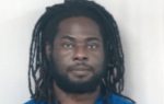 A man with long dreadlocks and a beard, wearing a blue shirt, looks directly at the camera against a plain, light background.