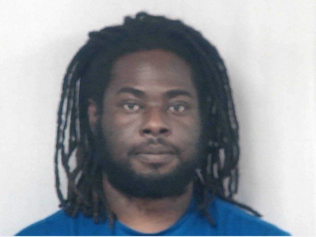 A man with long dreadlocks and a beard, wearing a blue shirt, looks directly at the camera against a plain, light background.