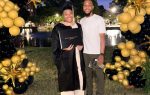 Graduate in cap and gown holding diploma, standing beside a man, under a black and gold balloon arch, outdoors in the evening.