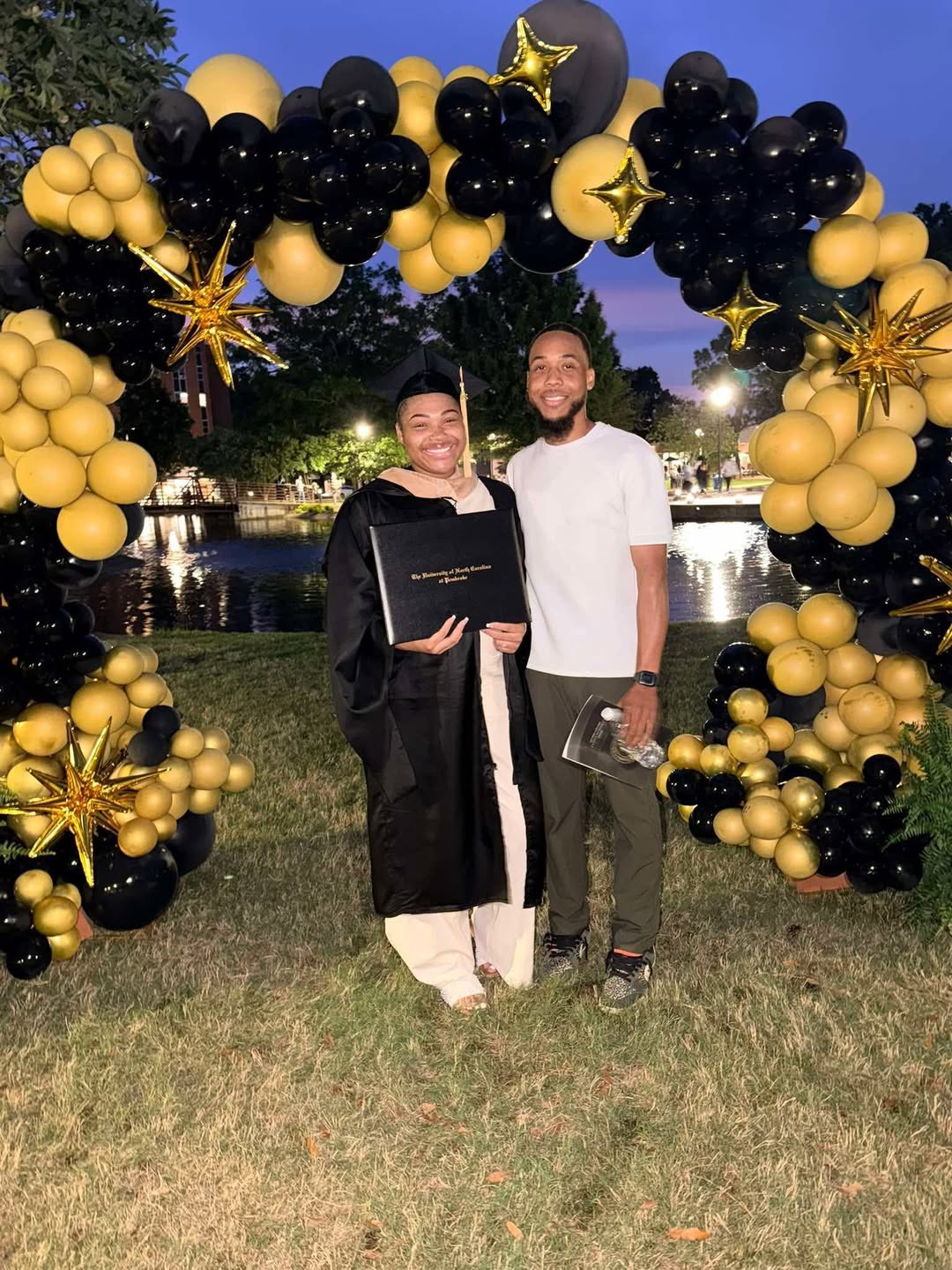 Graduate in cap and gown holding diploma, standing beside a man, under a black and gold balloon arch, outdoors in the evening.