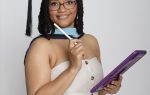 Woman in graduation cap and gown holding a tablet and stylus, smiling at the camera against a plain white background.