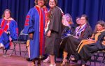 A group of people in academic regalia on stage, with one student and one faculty member smiling at the camera during a graduation ceremony.