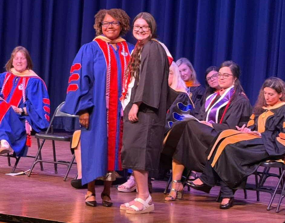 A group of people in academic regalia on stage, with one student and one faculty member smiling at the camera during a graduation ceremony.