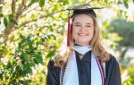 A smiling graduate in cap and gown stands outdoors among green trees, wearing a white stole and honor cords.