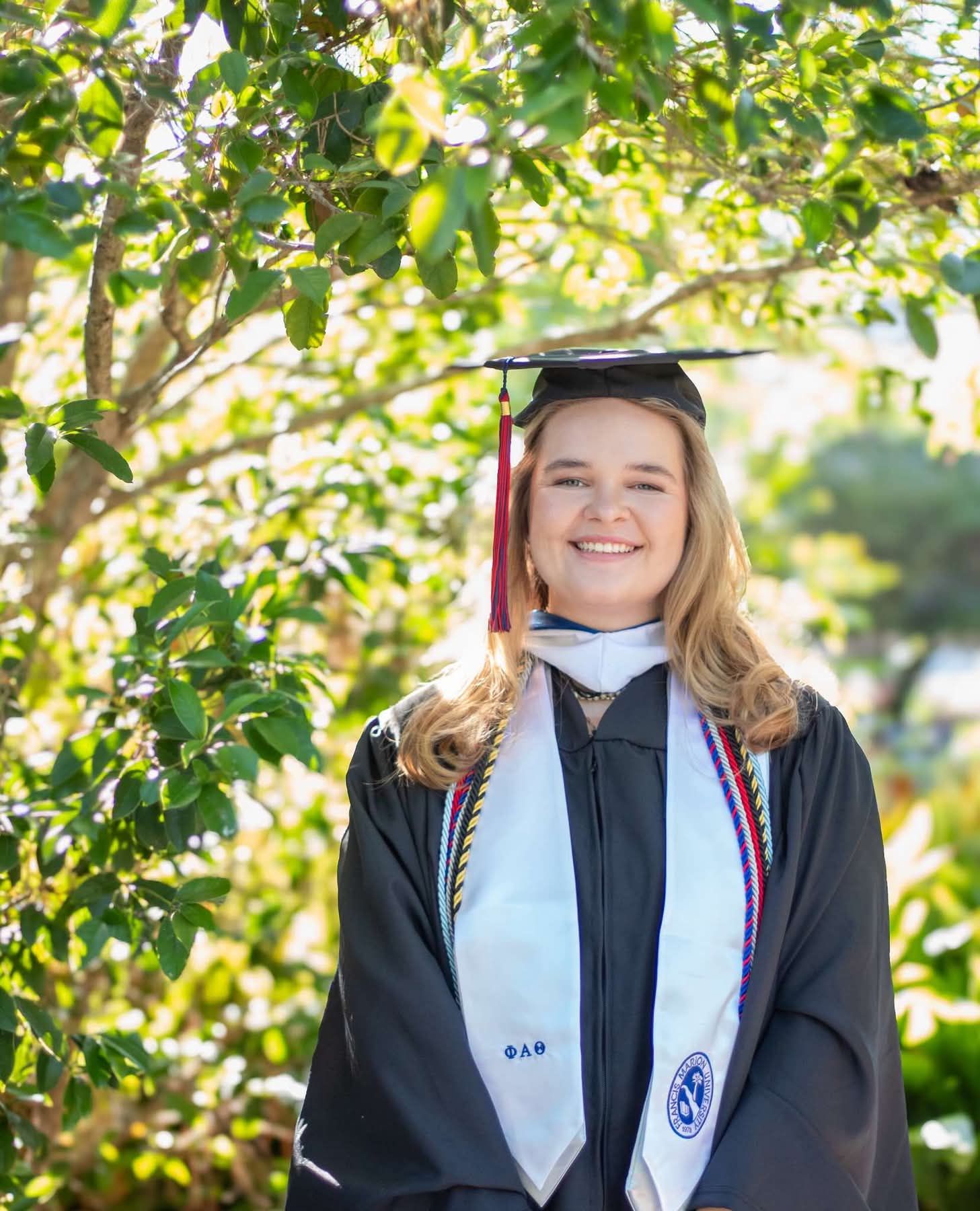 A smiling graduate in cap and gown stands outdoors among green trees, wearing a white stole and honor cords.