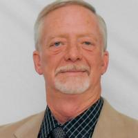 Older man with short gray hair, light beard, wearing a beige blazer over a checkered shirt and tie, posing against a plain light background.