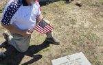 A man kneels at a gravesite holding an American flag, wearing a patriotic shirt and red beret, honoring a fallen soldier named Anthony Chad Owens.