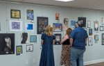 Three people observe colorful artwork displayed on a gallery wall, with one person pointing at a specific piece.