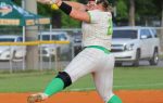 A softball pitcher in mid-throw on a dirt field, wearing a white and green uniform, with cars and trees in the background.