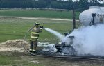 Firefighter spraying water on a smoking, burned vehicle near a jet fuel tank in an open grassy area with trees in the background.