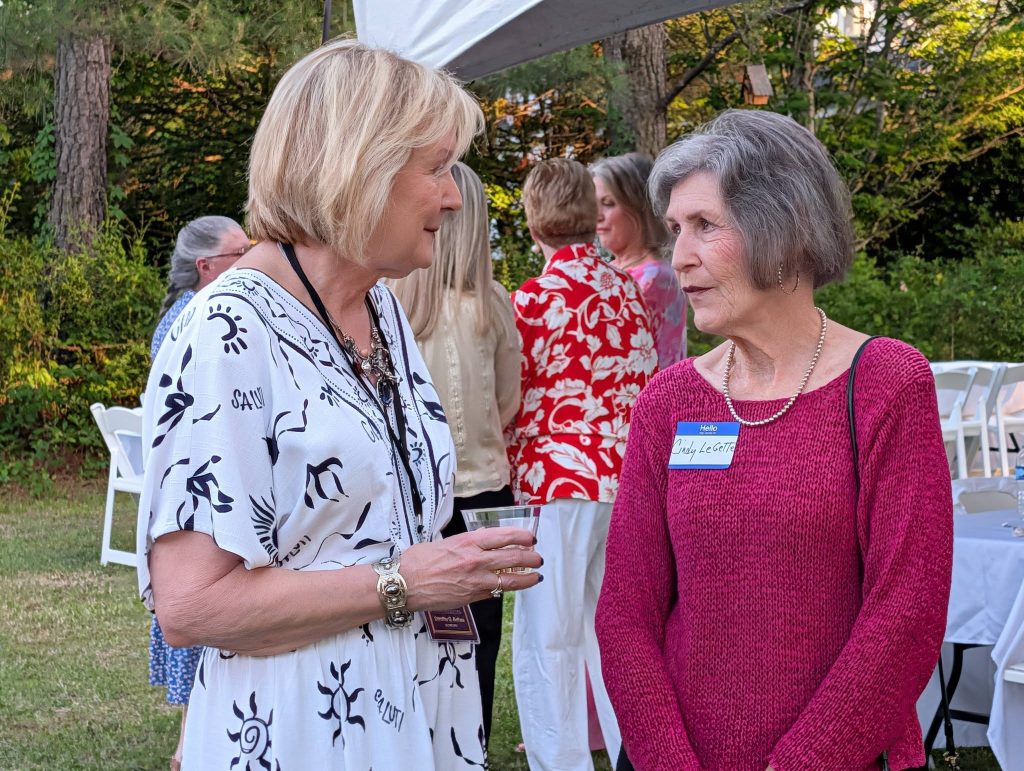 Two women have a conversation at an outdoor event, with other people and white chairs visible in the background. One woman wears a nametag.