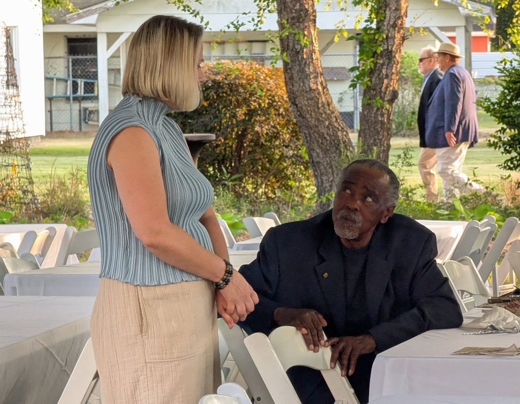 A woman stands and talks to a seated man at an outdoor event with white chairs and tables, while two men walk in the background.