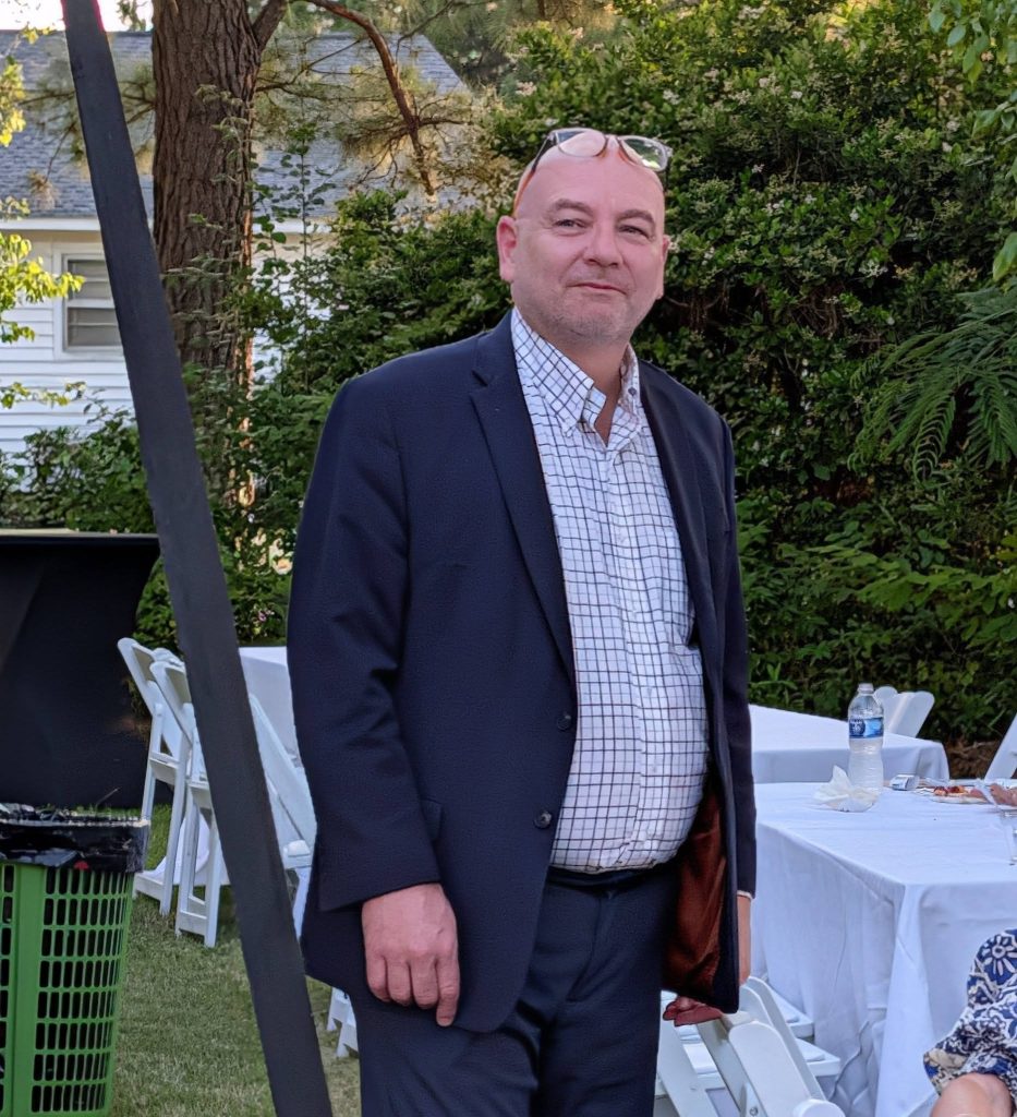 Man in suit and checked shirt stands outdoors at an event with white tables, chairs, and greenery in the background.