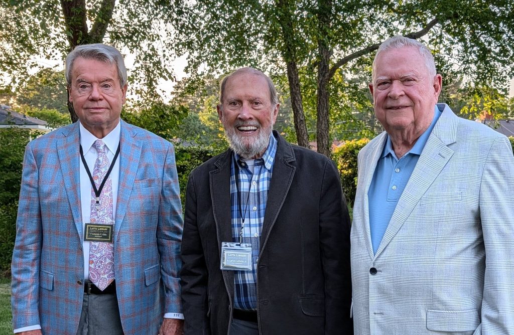 Three older men wearing jackets and name tags stand outdoors in front of green trees, smiling for the camera.