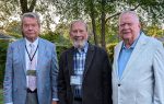 Three older men wearing jackets and name tags stand outdoors in front of green trees, smiling for the camera.