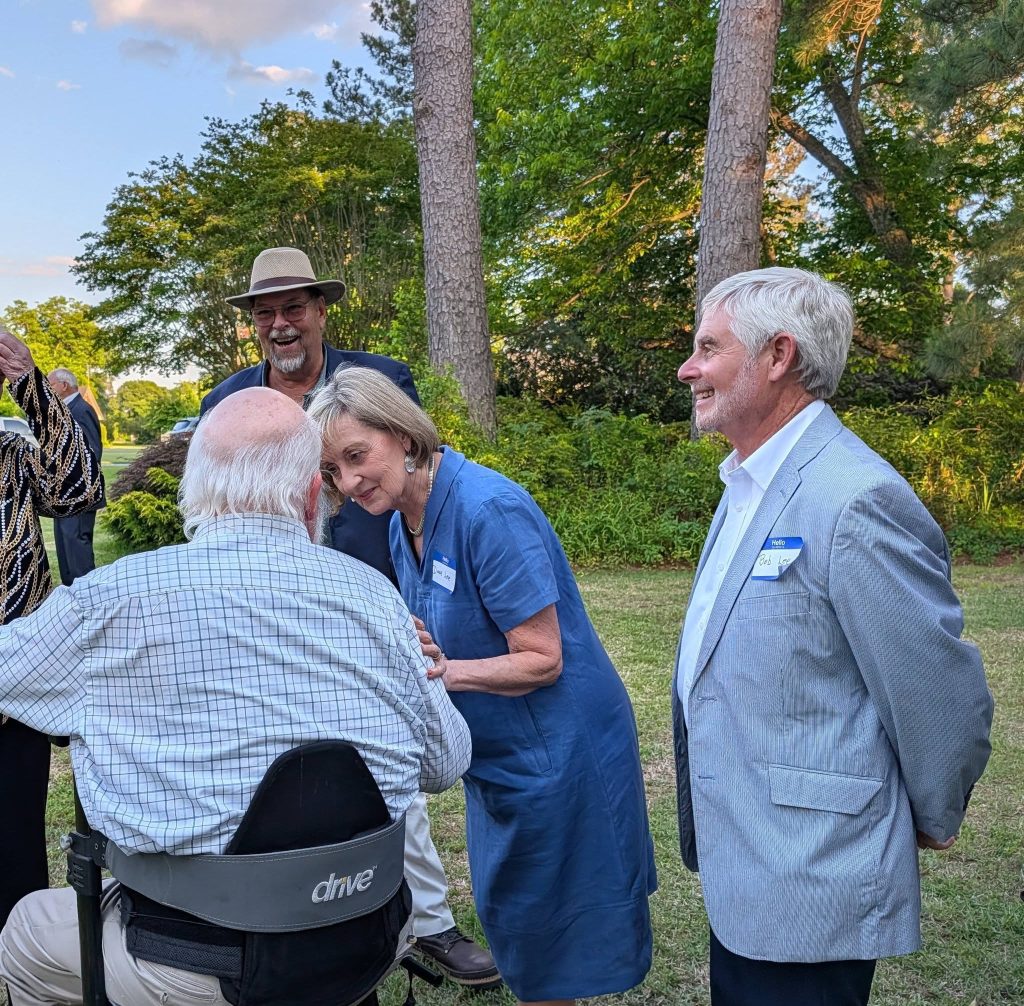 A group of older adults socializing outdoors in a garden, with one person in a wheelchair and others smiling and conversing around them.