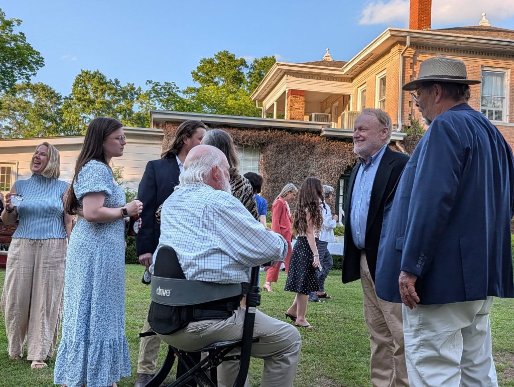 People socializing outdoors at a garden party, some standing, one man seated in a wheelchair, with a large house in the background on a sunny day.