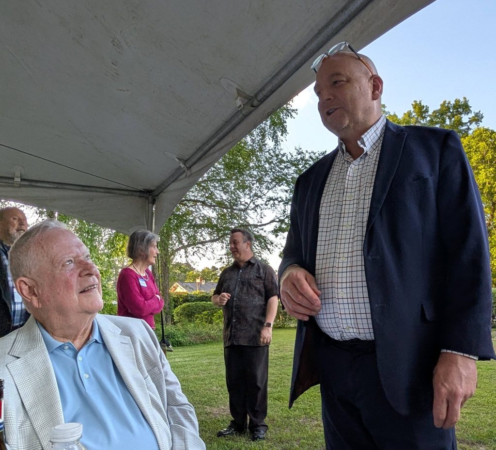 Two men in suits converse under a tent at an outdoor event while others talk in the background, surrounded by trees and greenery.