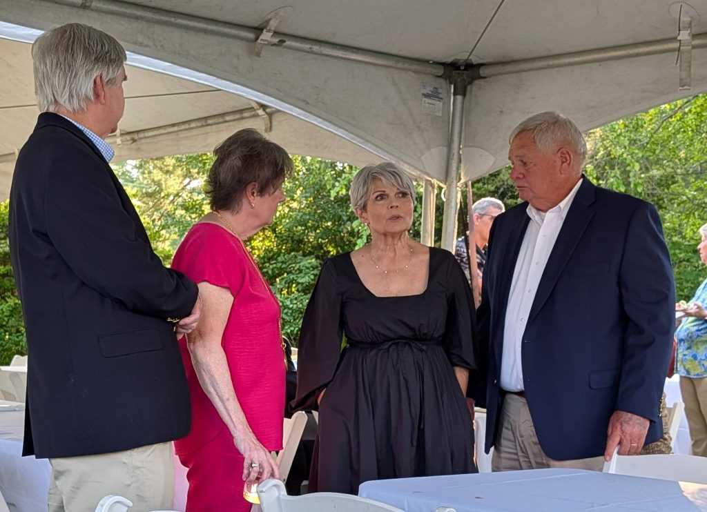 Four adults, two men and two women, stand and talk under a canopy at an outdoor event with white chairs and tables.