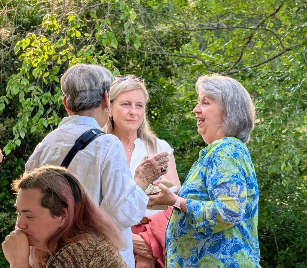 Three women converse outdoors among green trees, one holding a plate and fork, while another person sits in the foreground, appearing thoughtful.