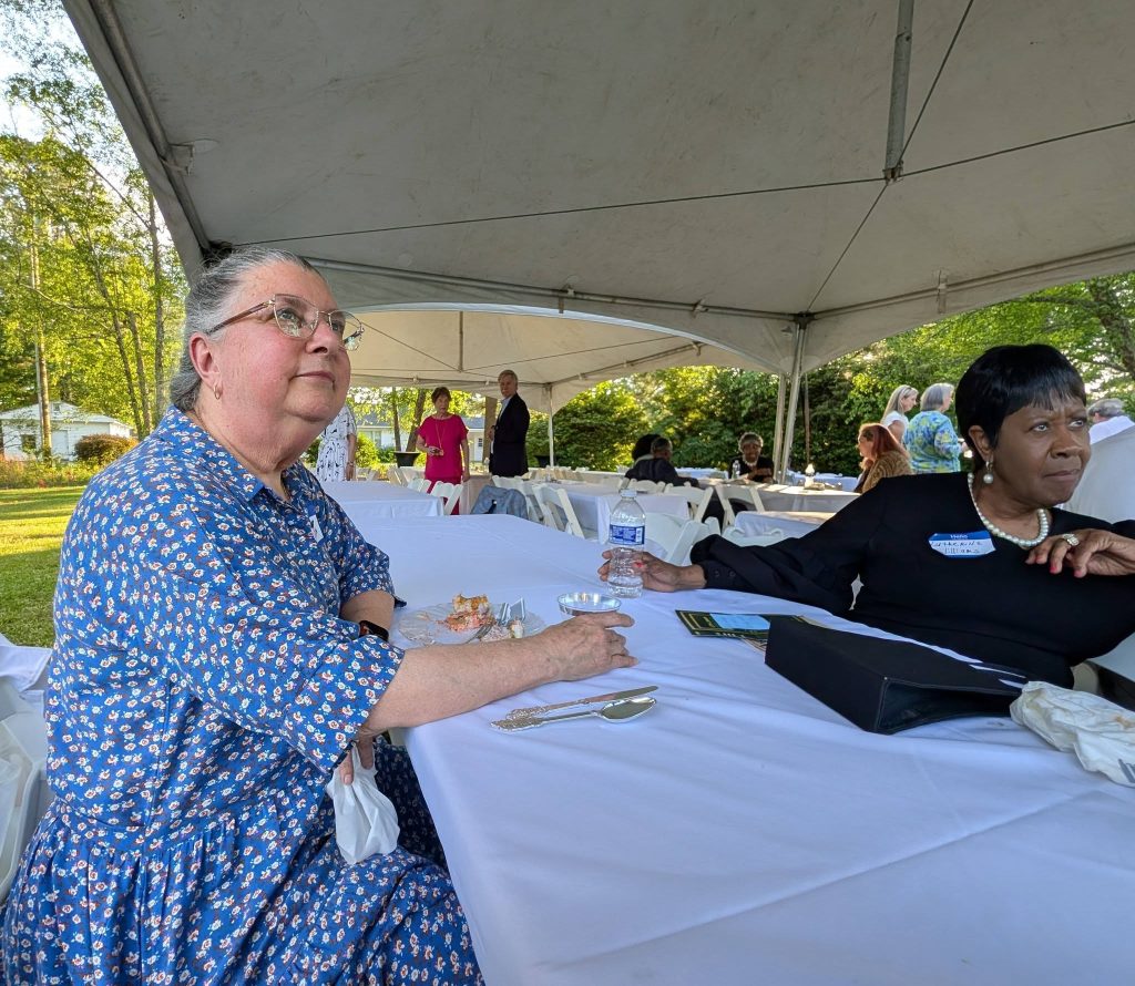Two women sit at a table under a large outdoor tent during a daytime event, with people and trees visible in the background.