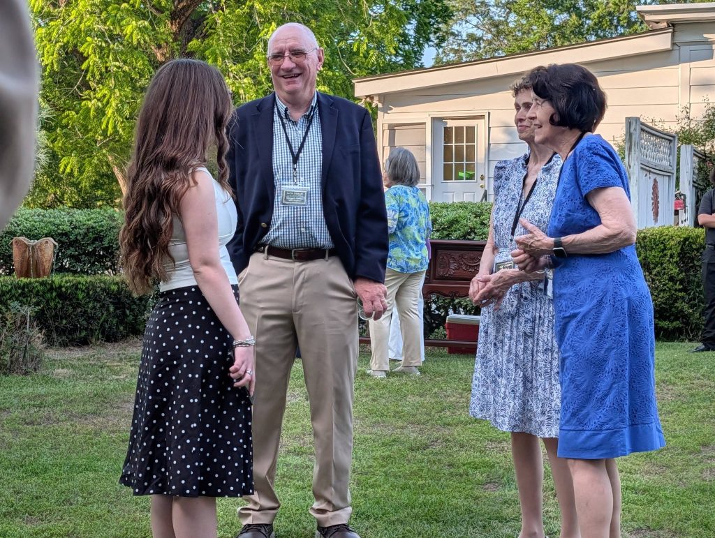 Four adults stand outdoors on grass, smiling and conversing in front of a house, surrounded by greenery and trees on a sunny day.
