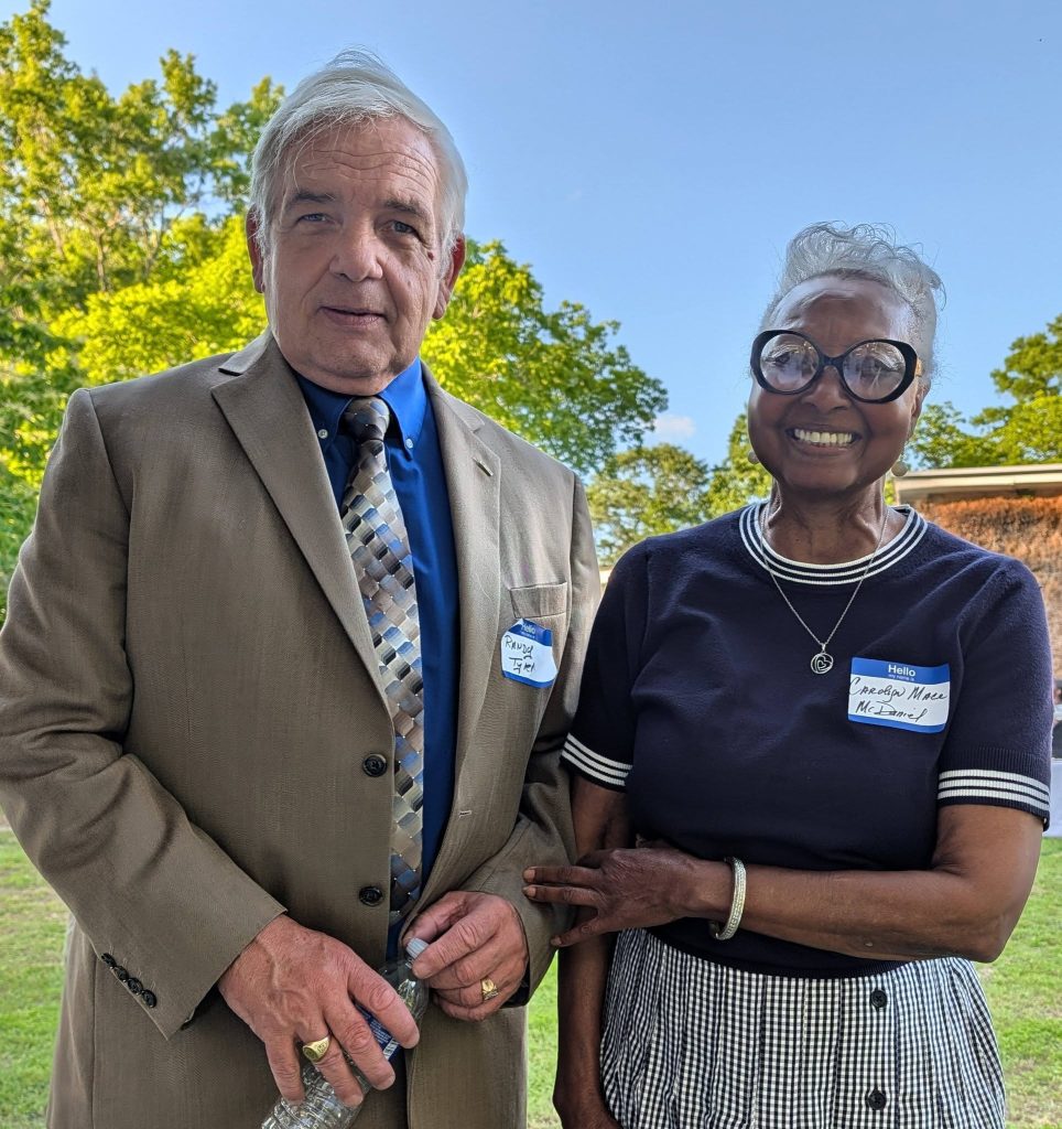 elderly man in a suit and an elderly woman in glasses are smiling outdoors, both wearing nametags, with trees and blue sky in the background.