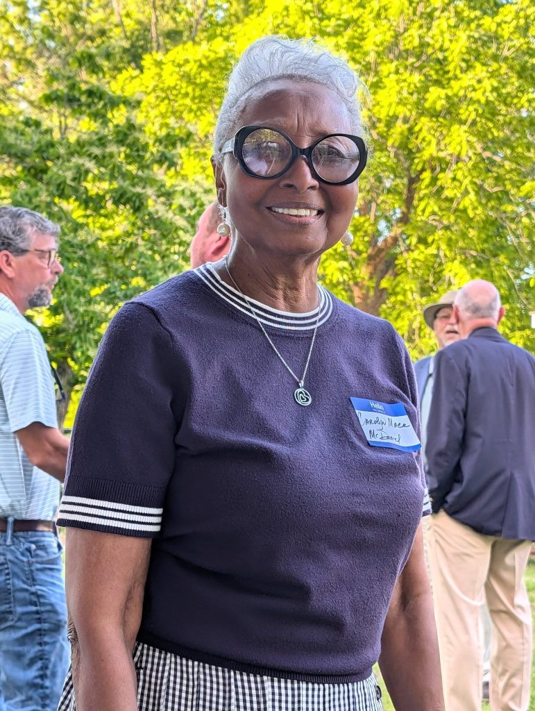 Smiling woman with short gray hair, large glasses, and a name tag, standing outdoors among people with green trees in the background.
