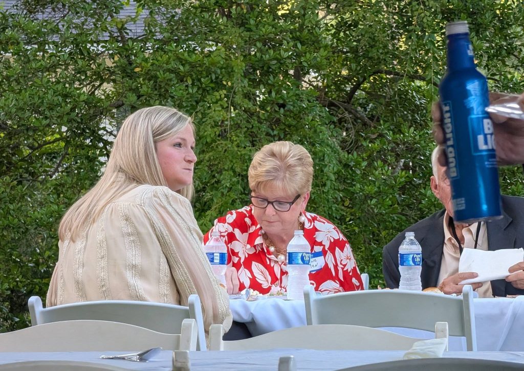 Three people sit at an outdoor table with water bottles and food, while someone holds up a blue Bud Light bottle in the foreground.