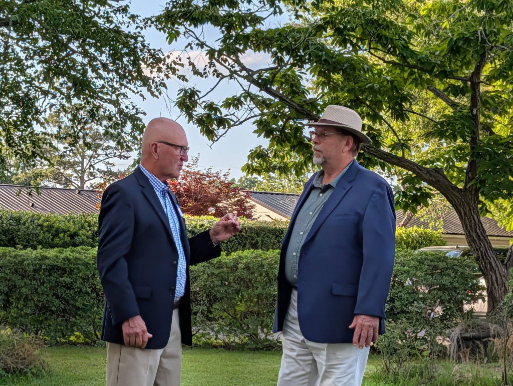 Two men in blazers talk outdoors near trees and bushes; one is wearing a hat, the other glasses, with a house and greenery in the background.