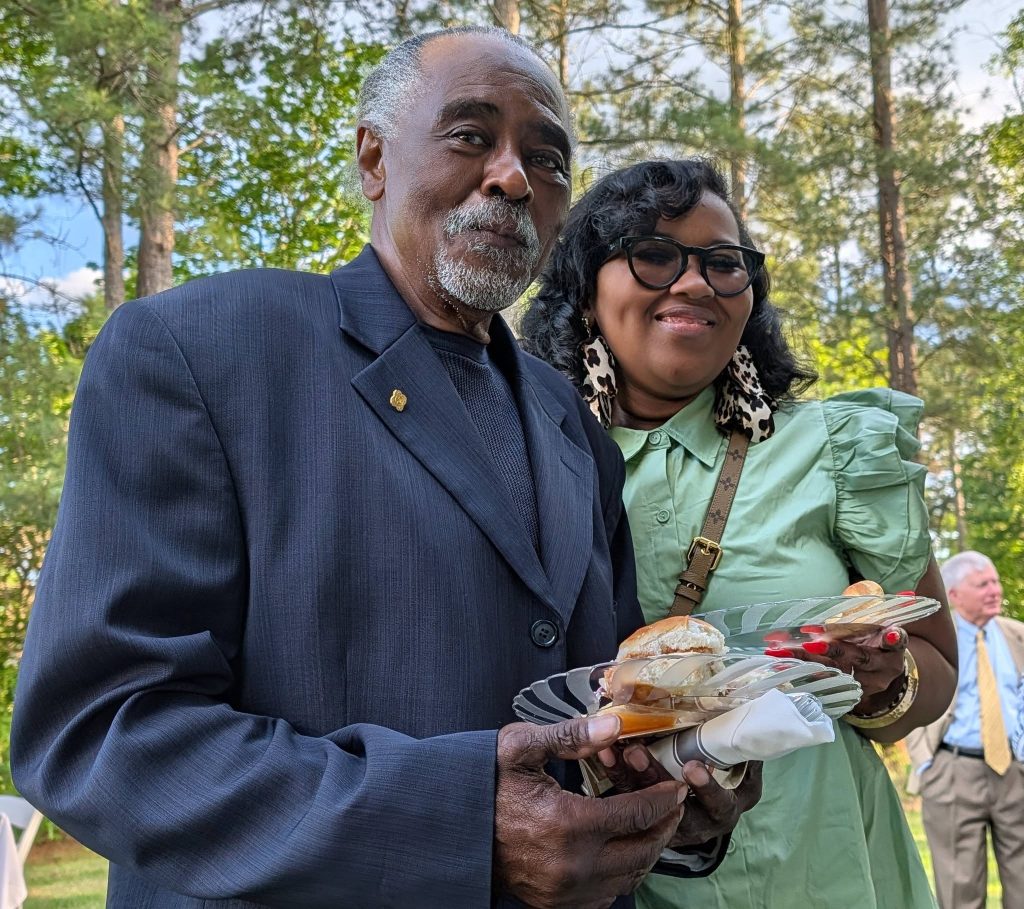 older man in a suit and a woman in glasses hold plates of food, smiling at an outdoor event with trees and people in the background.
