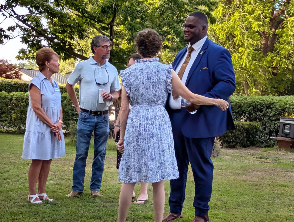 Four adults stand outside on grass, engaged in conversation. One man is in a blue suit, the others are casually dressed. Green trees and bushes are in the background.