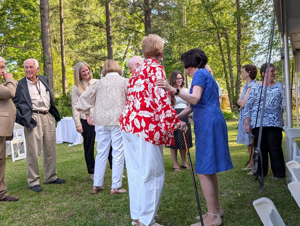 A group of adults chatting outdoors on grass, surrounded by trees, dressed in semi-formal attire, with white chairs and a tent nearby.